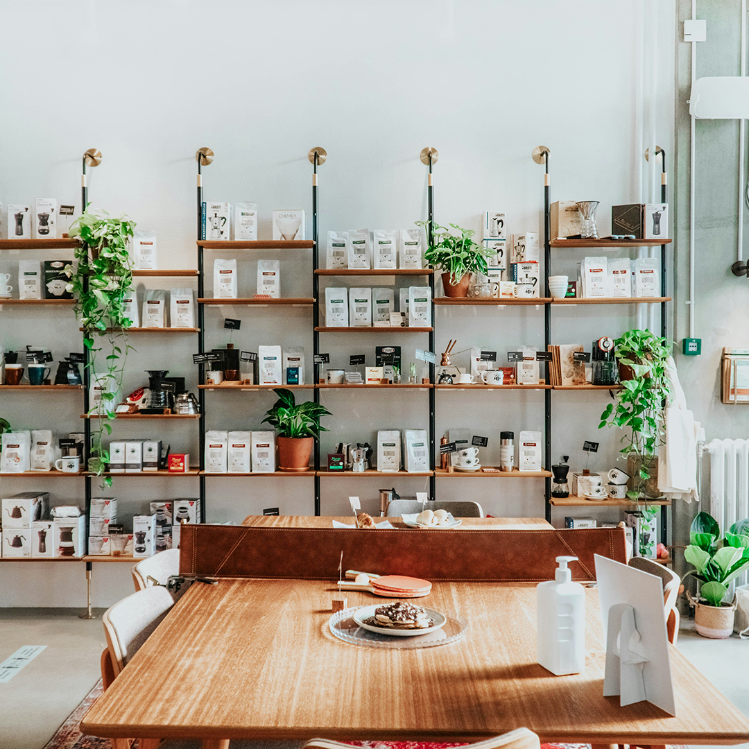 étagères en bois remplies de paquets de café et de produits fins dans un café-boutique lumineux, avec plantes décoratives et grande table en bois au premier plan