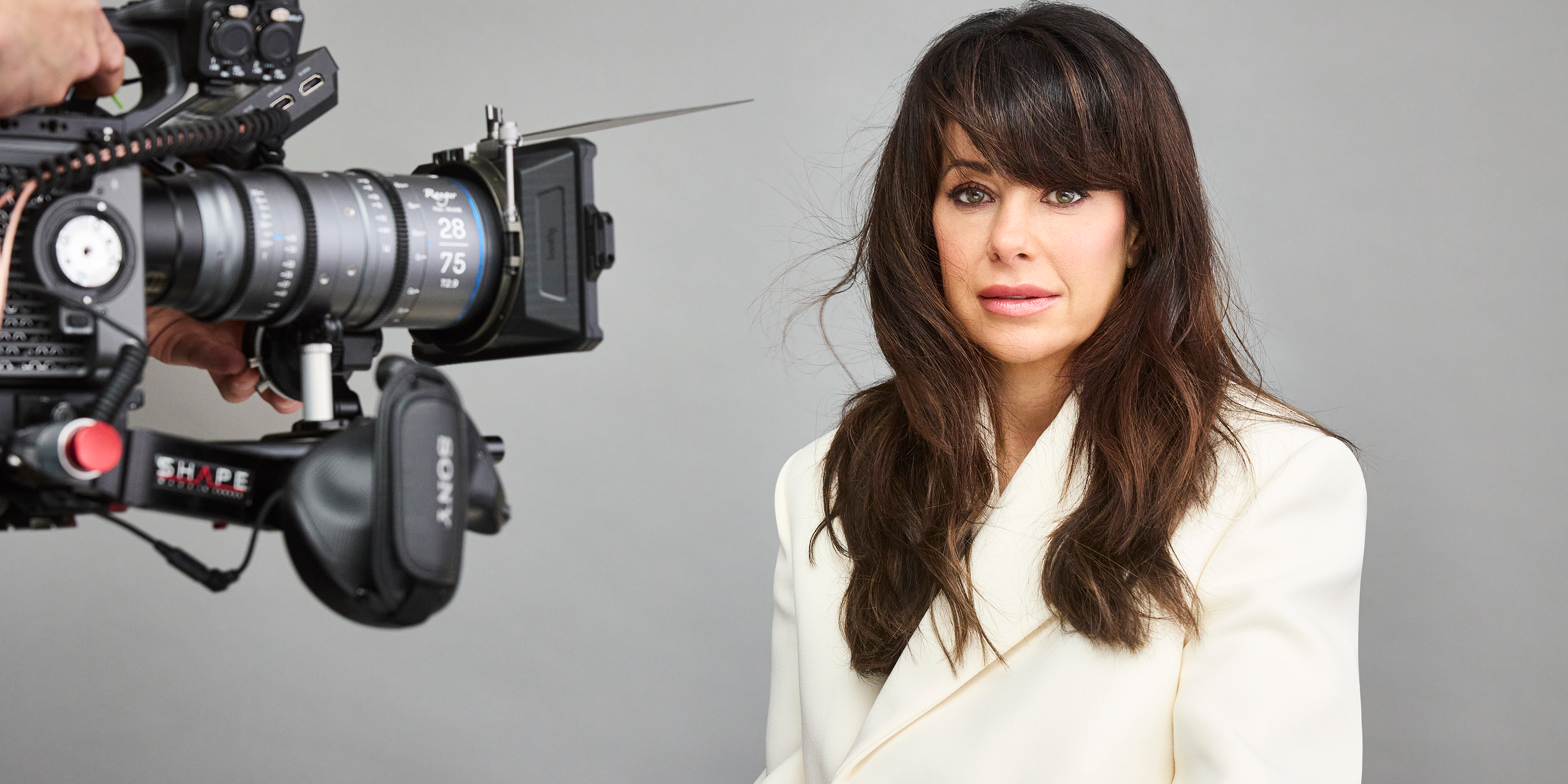 Marie-Claude_Savard en veste blanche assise devant une caméra de tournage professionnelle, sur fond gris
