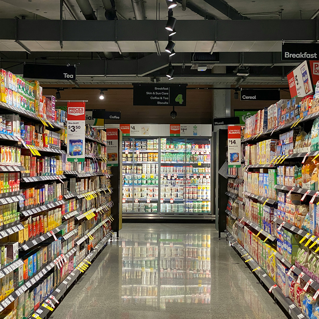 allée d’un supermarché avec des étagères remplies de céréales, biscuits, thés et cafés, menant vers une section réfrigérée éclairée