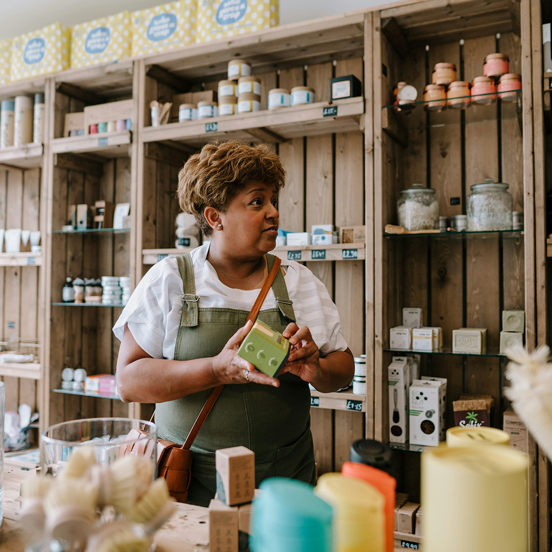 femme dans une boutique artisanale tenant un produit vert, entourée d’étagères en bois remplies de savons, bocaux et cosmétiques naturels